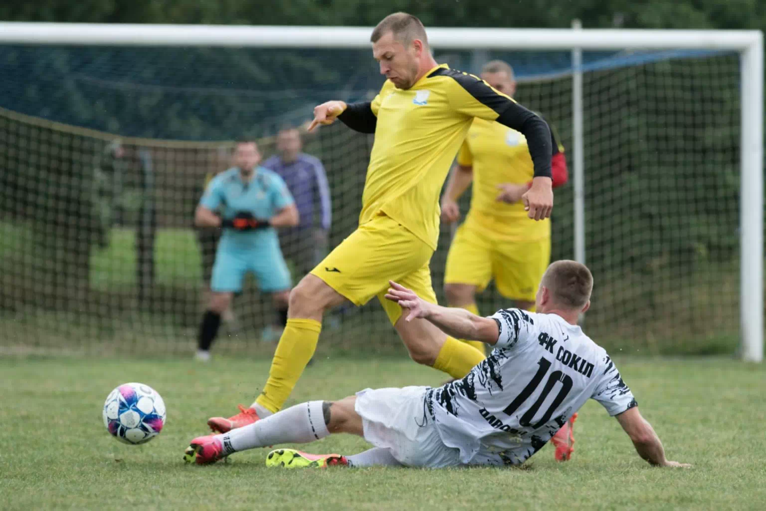 Технічний момент на футбольному полі: гравець у жовтій формі намагається зіграти м’ячем біля воріт, тоді як суперник у білій формі з номером 10 виконує підкат, намагаючись відібрати м’яч; на задньому плані видно воротаря та сітку воріт. ⚽